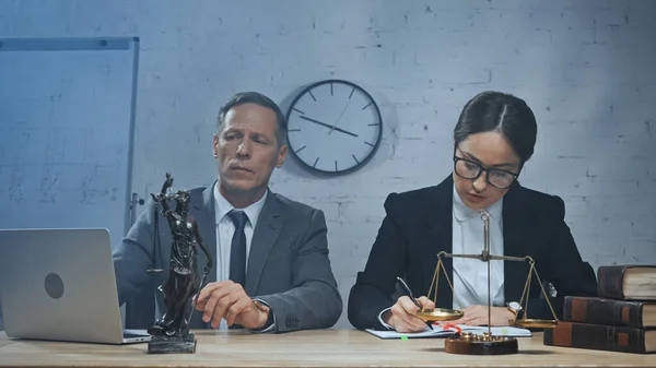 Insurance agent writing on notebook near colleague using laptop and books on table — Stock Photo