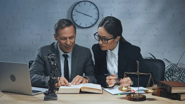 Улыбающийся страховой агент читает книгу рядом с коллегой с ручкой и ноутбуком на столе — стоковое фото
