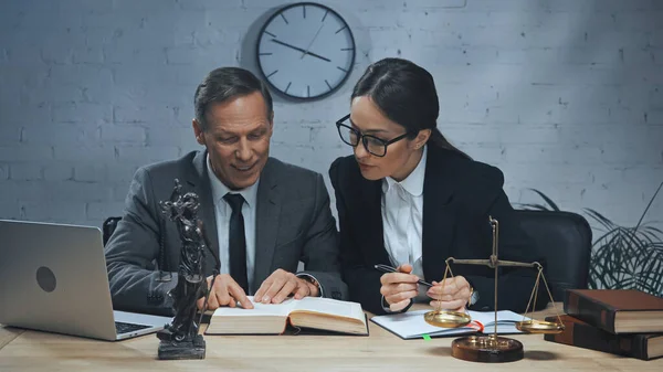 Agente de seguros sonriente leyendo libro cerca de colega con la pluma y el ordenador portátil en la mesa - foto de stock