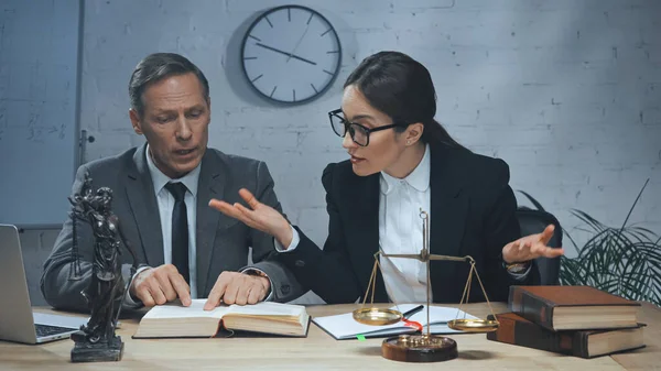 Insurance agent showing shrug gesture near colleague reading book, laptop and scales in office — Stock Photo