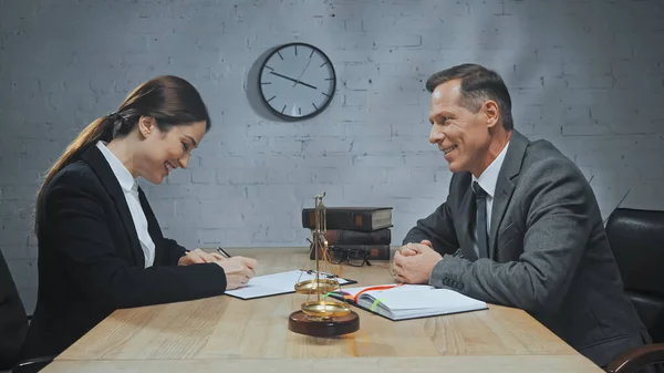 Alegre agente de seguros escribiendo en el documento cerca de colega, escalas y cuaderno — Stock Photo