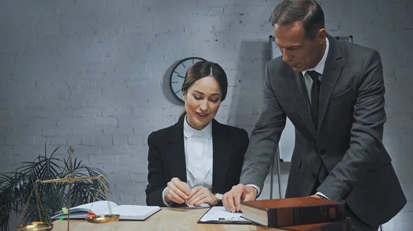 Smiling insurance agent sitting near colleague, clipboard and books on blurred foreground — Stock Photo