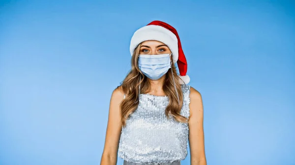 Жінка в медичній масці і капелюсі Санта дивиться на камеру ізольовано на синьому — стокове фото