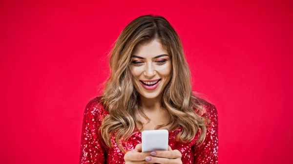 Mujer asombrada con mensajes de texto de pelo ondulado en el teléfono inteligente aislado en rojo - foto de stock