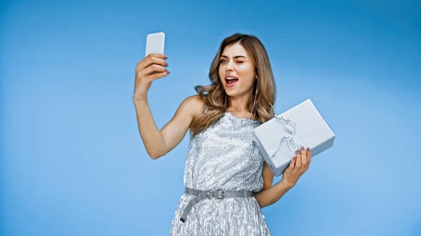 Verblüffte Frau zwinkert, hält Geschenkbox und macht Selfie auf blau — Stockfoto