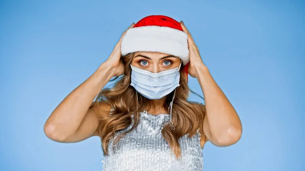 Жінка з хвилястим волоссям в медичній масці в капелюсі Санта ізольована на синьому — стокове фото