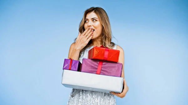 Happy woman covering mouth while holding wrapped presents isolated on blue — Stock Photo