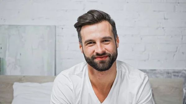 Joyful bearded man smiling at camera while sitting in bedroom — Stock Photo