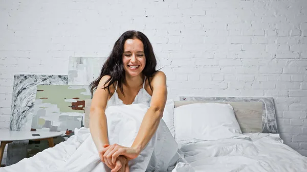 Fröhliche brünette Frau mit geschlossenen Augen dehnt sich, während sie im Bett sitzt — Stockfoto