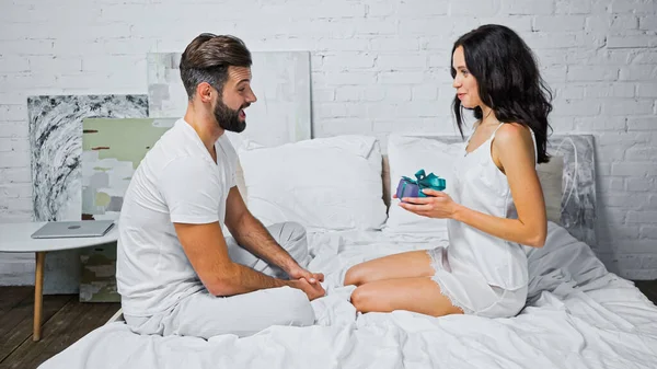 Vue latérale de la femme brune souriante tenant boîte cadeau près de l'homme excité dans la chambre — Photo de stock