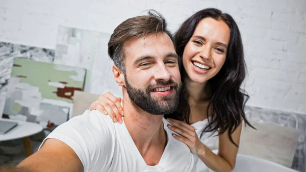 Hombre barbudo feliz y mujer morena sonriendo a la cámara en el dormitorio - foto de stock