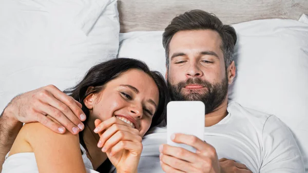 Man hugging cheerful girlfriend while using smartphone in bed — Stock Photo