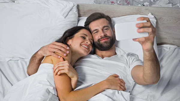 Felice uomo barbuto abbracciando fidanzata allegra mentre prende selfie a letto — Foto stock
