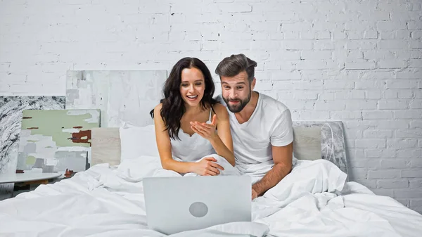 Mulher animado apontando com a mão para laptop enquanto assiste filme com namorado na cama — Fotografia de Stock