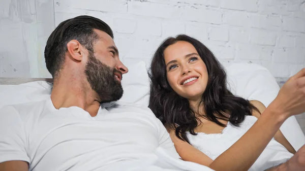 Mulher morena feliz falando com namorado sorridente enquanto deitado na cama — Fotografia de Stock