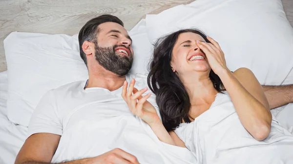 Rindo mulher tocando rosto enquanto deitado na cama perto de homem feliz — Fotografia de Stock