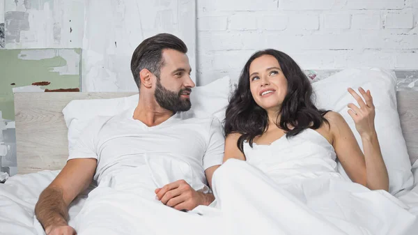 Sorrindo morena mulher gesticulando enquanto conversa com namorado barbudo na cama — Fotografia de Stock