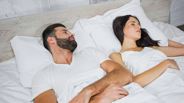 Bearded man looking at brunette woman sleeping nearby in bed — Stock Photo