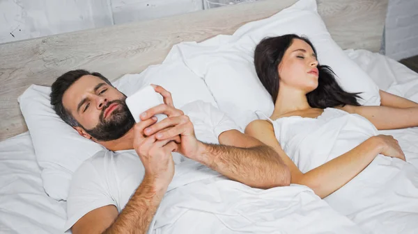 Bearded man chatting on mobile phone near sleeping girlfriend in bed — Stock Photo