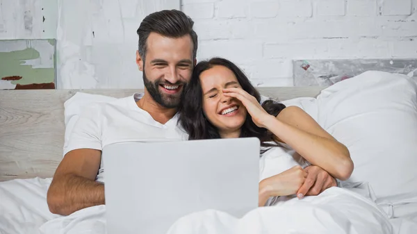 Laughing woman touching face while watching movie on laptop with cheerful boyfriend in bed — Stock Photo