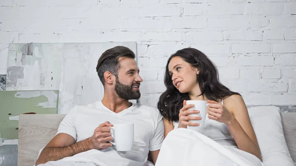 Счастливая молодая пара с чашками кофе говорить в постели — стоковое фото