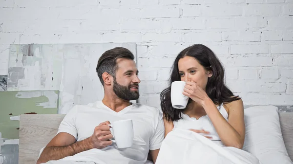 Happy bearded man looking at girlfriend drinking coffee in bed — Stock Photo