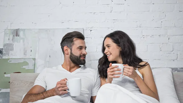 Cheerful man and woman looking at each other while holding cups of coffee in bed — Stock Photo