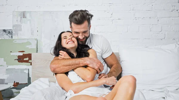 Excited woman with closed eyes laughing while cheerful boyfriend embracing her in bedroom — Stock Photo