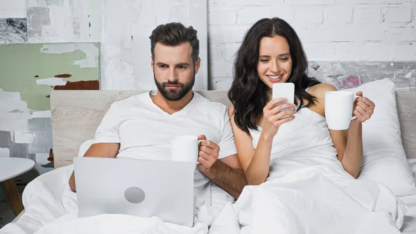 Sonriente mujer charlando en el teléfono inteligente cerca de hombre utilizando el ordenador portátil en la cama - foto de stock