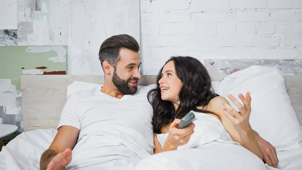 Cheerful couple looking at each other while watching tv in bed — Stock Photo