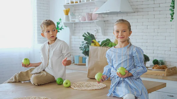 Улыбающаяся девушка с яблоками сидит на кухонном столе рядом с братом и бумажным пакетом — стоковое фото
