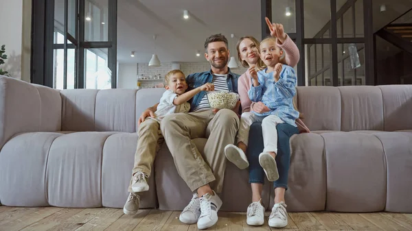 Smiling woman pointing with finger near children and husband with popcorn at home — Stock Photo