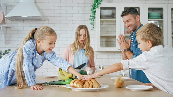 Enfants prenant des croissants près des parents au premier plan flou dans la cuisine — Photo de stock