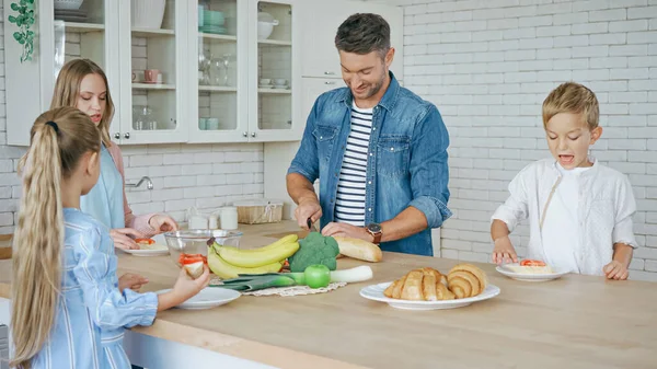 Sorridente padre taglio baguette vicino alla famiglia in cucina — Foto stock