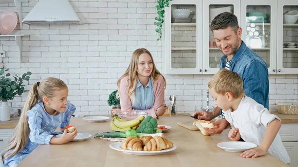 Smiling man giving baguette to son near family in kitchen — Stock Photo