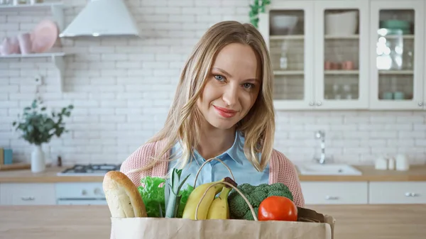 Smiling woman with fresh vegetables and baguette in paper bag in kitchen — Stock Photo