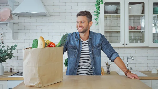 Hombre sonriendo cerca de bolsa de papel con comida en la mesa de la cocina - foto de stock
