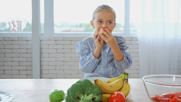 Girl eating baguette near fresh vegetables and fruits on blurred foreground — Stock Photo
