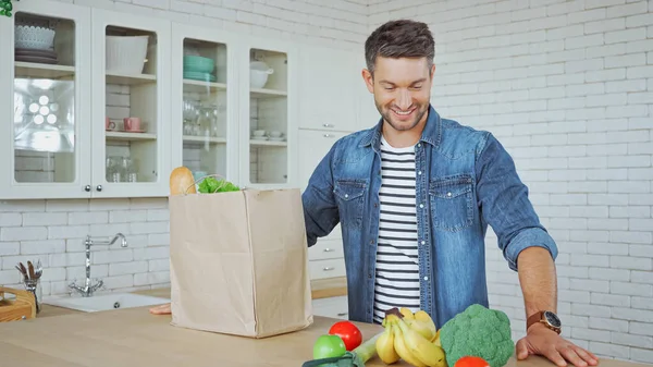 Smiling man looking at fresh food near paper package on kitchen table — Stock Photo