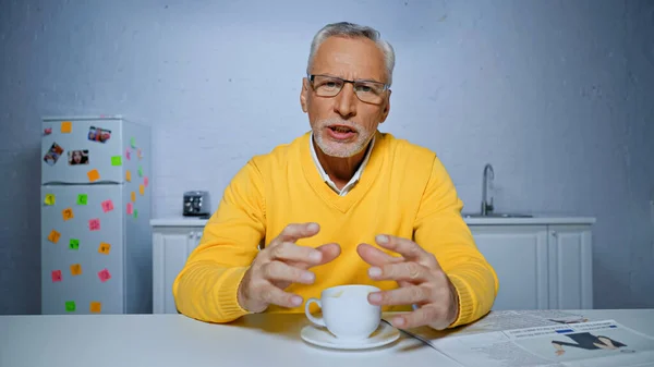 Senior man in eyeglasses talking at camera near cup during video chat — Stock Photo