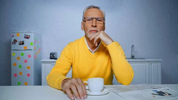 Uomo anziano in occhiali guardando la fotocamera vicino a tazza di caffè e giornale in primo piano sfocato — Foto stock