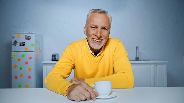 Allegro uomo anziano guardando la fotocamera vicino alla tazza di caffè in cucina — Foto stock