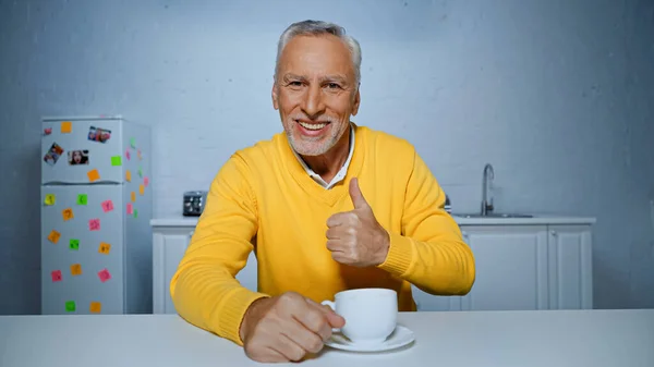 Smiling senior man showing like near cup of coffee in kitchen — Stock Photo