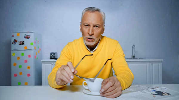 Пожилой мужчина, держащий очки во время видеочата на кухне — стоковое фото