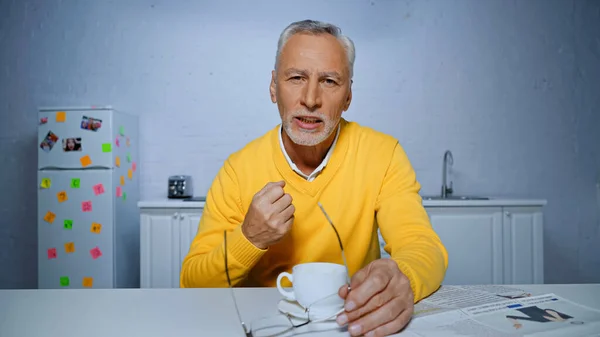 Elderly man holding eyeglasses and showing yeah gesture near cup during video call — Stock Photo