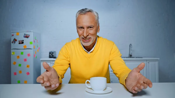 Улыбающийся мужчина, указывающий руками на камеру возле чашки на кухонном столе — стоковое фото