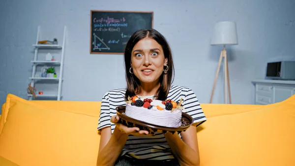 Aufgeregte Frau blickt in die Kamera, während sie Kuchen auf gelbem Sofa hält — Stockfoto