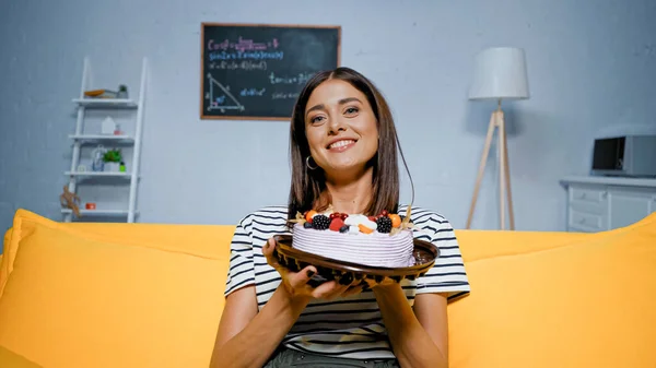 Mulher feliz mostrando bolo saboroso com bagas na câmera na sala de estar — Fotografia de Stock