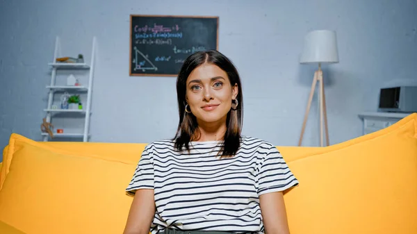 Улыбающаяся женщина в полосатой футболке смотрит в камеру на диване дома — стоковое фото