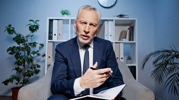 Älterer Geschäftsmann im Anzug hält Stift und Notizbuch in der Hand, während er im Büro vor der Kamera spricht — Stockfoto